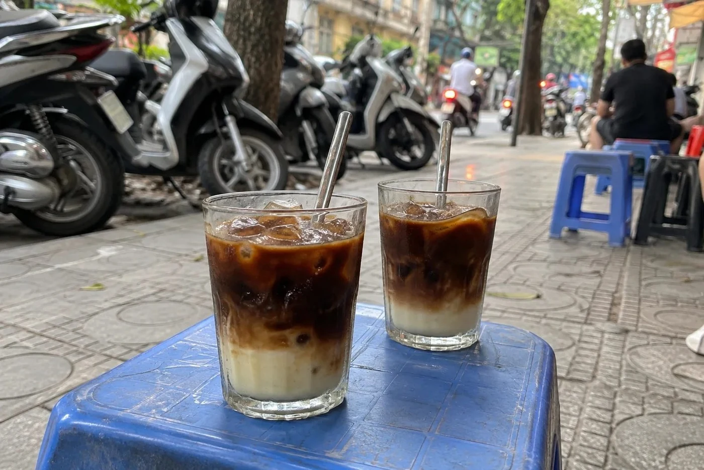 Two iced coffees on a blue plastic stool on a Vietnamese pavement with parked scooters