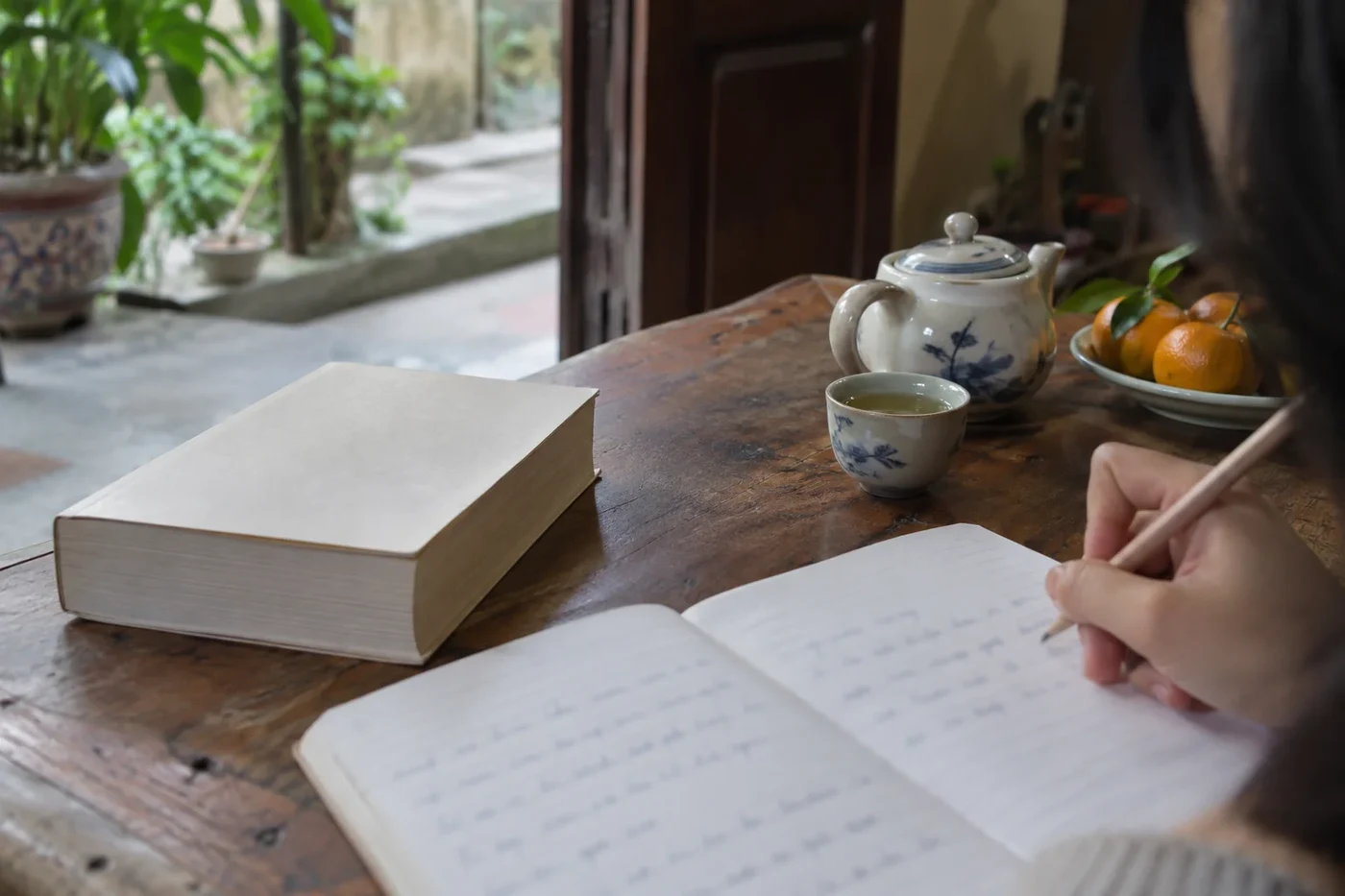 Hand writing in a notebook beside a cup of tea and a book on a wooden table