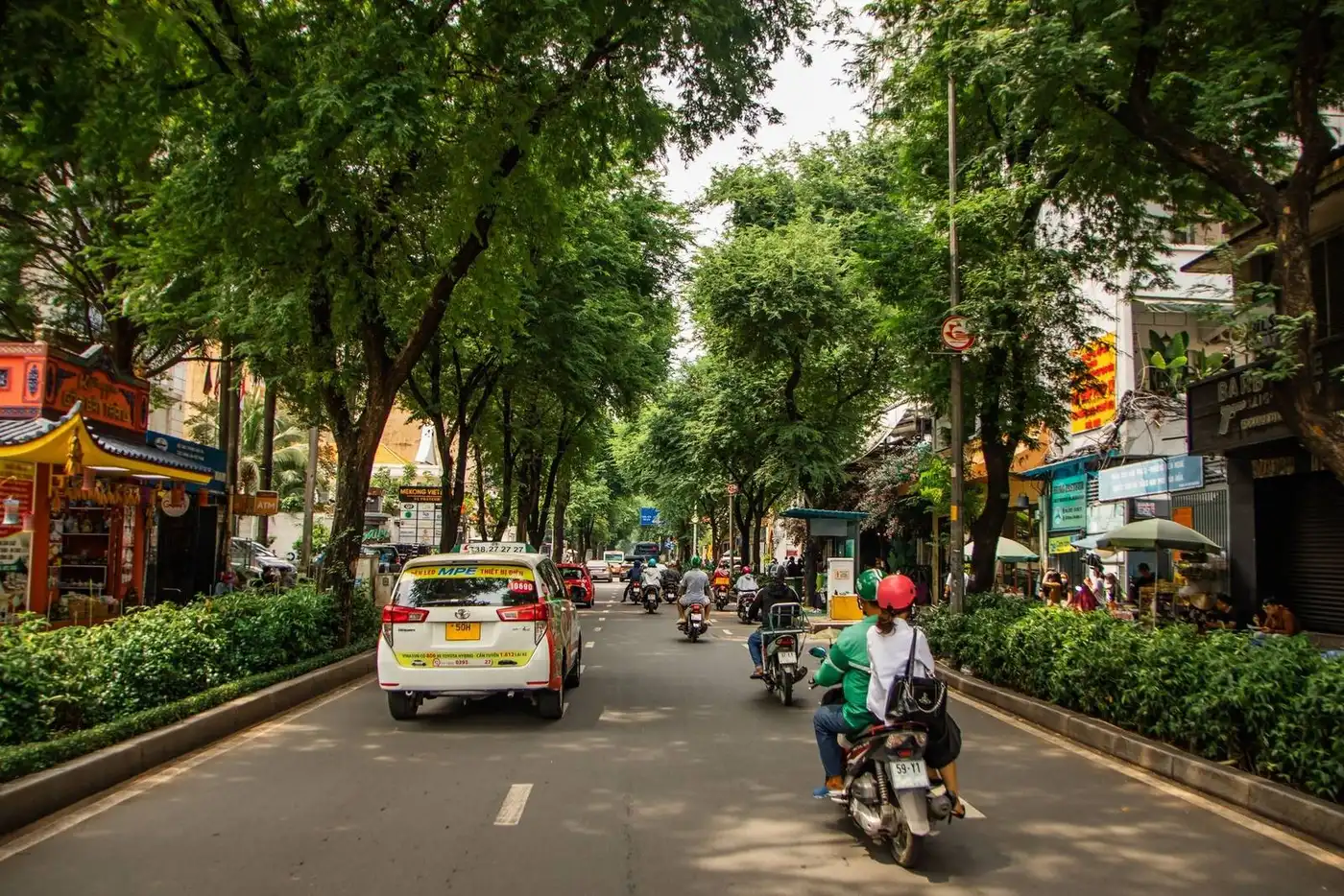 Tree-lined city street with taxis, motorbikes, and an ATM sign