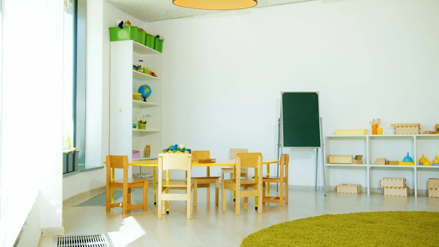 Bright classroom with a round yellow table, child-size wooden chairs, toy shelves and a small blackboard