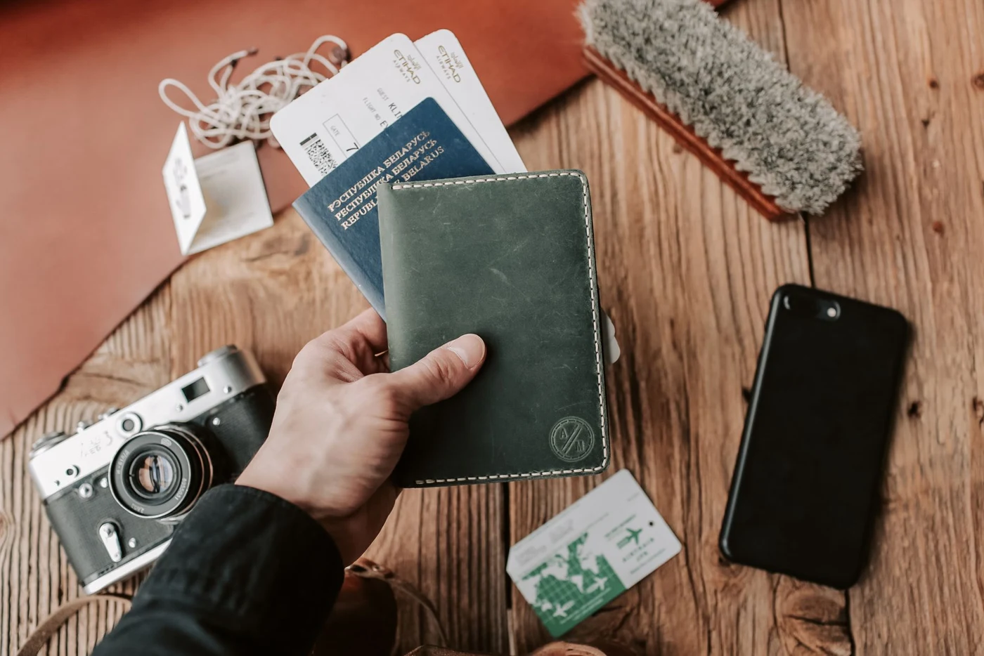A hand holding a dark blue passport with boarding passes next to a black smartphone and a film camera on a wooden table