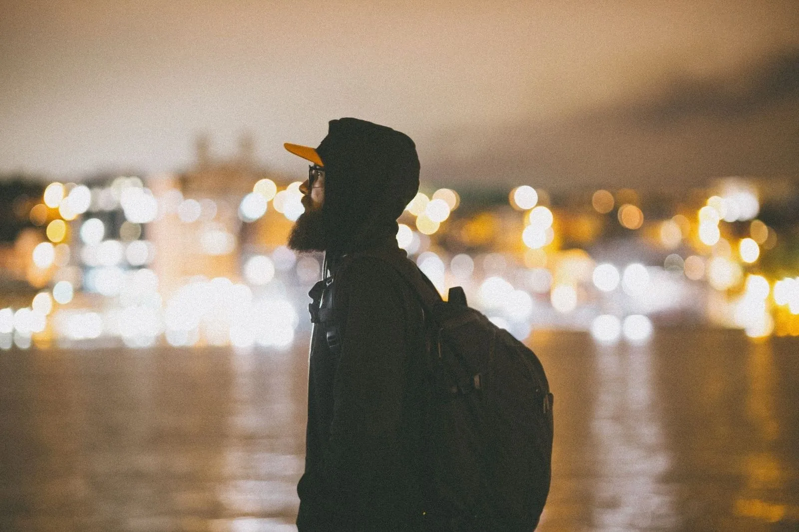 Backpacker standing alone by a river at night with city lights