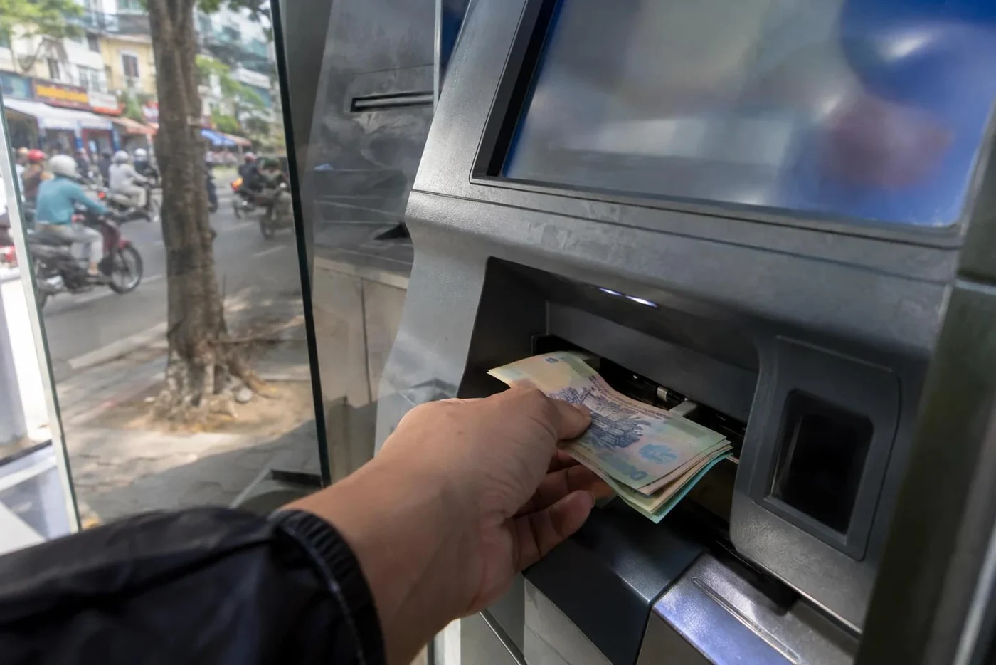 Hand taking dong banknotes from a street ATM slot with motorbikes behind