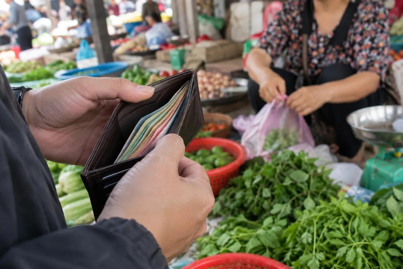 Hands holding a wallet full of dong banknotes at a vegetable stall