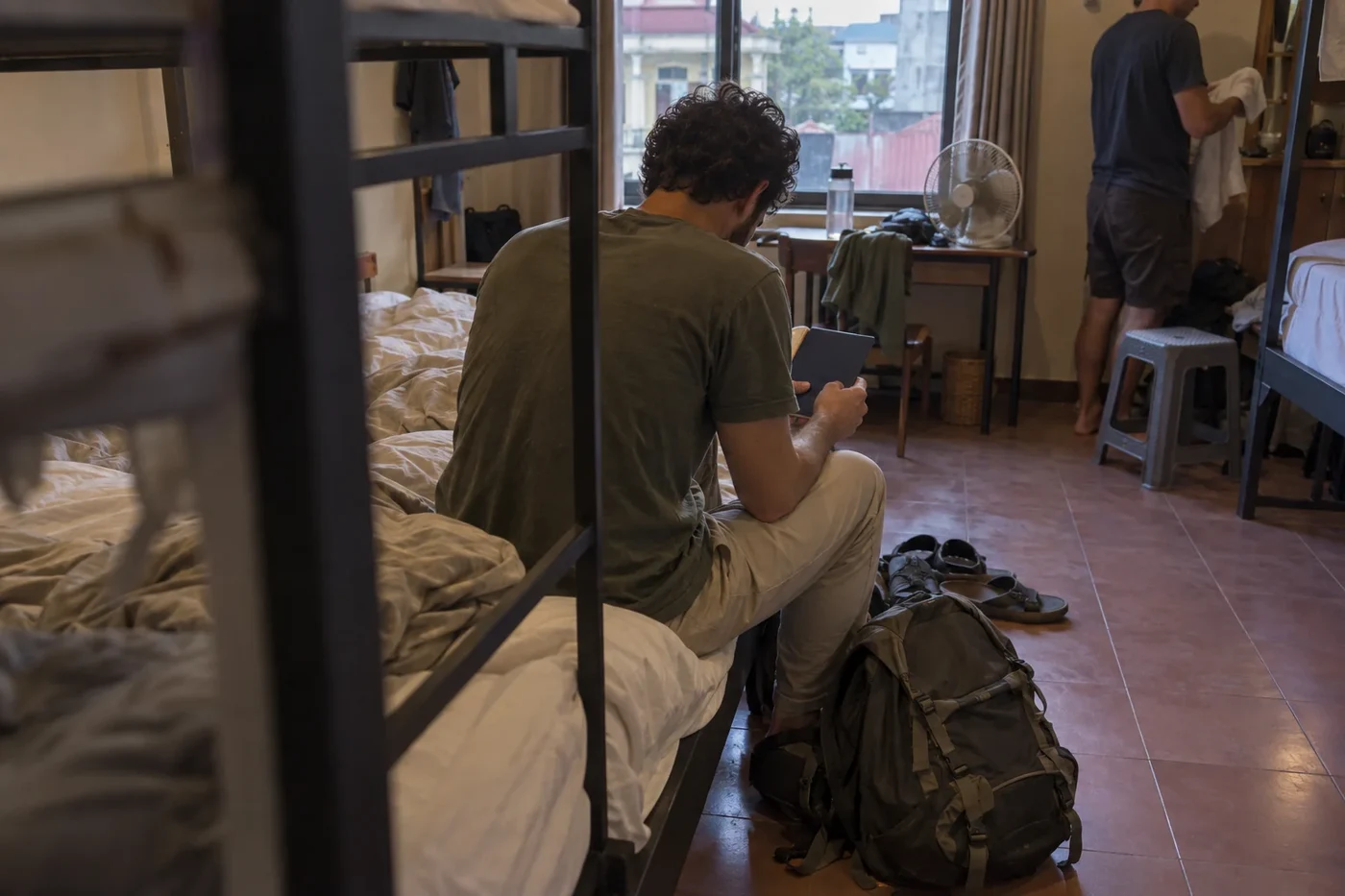 A traveller sitting on a bed checks his phone, a backpack on the floor
