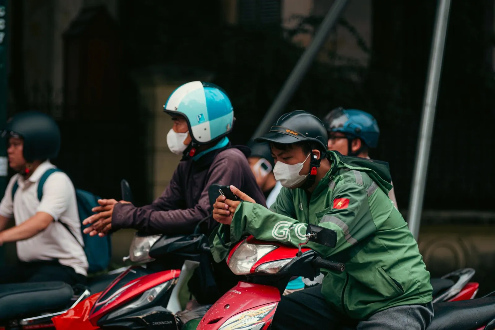 Motorbike riders in helmets and masks waiting in traffic, one checking a phone, in Vietnam