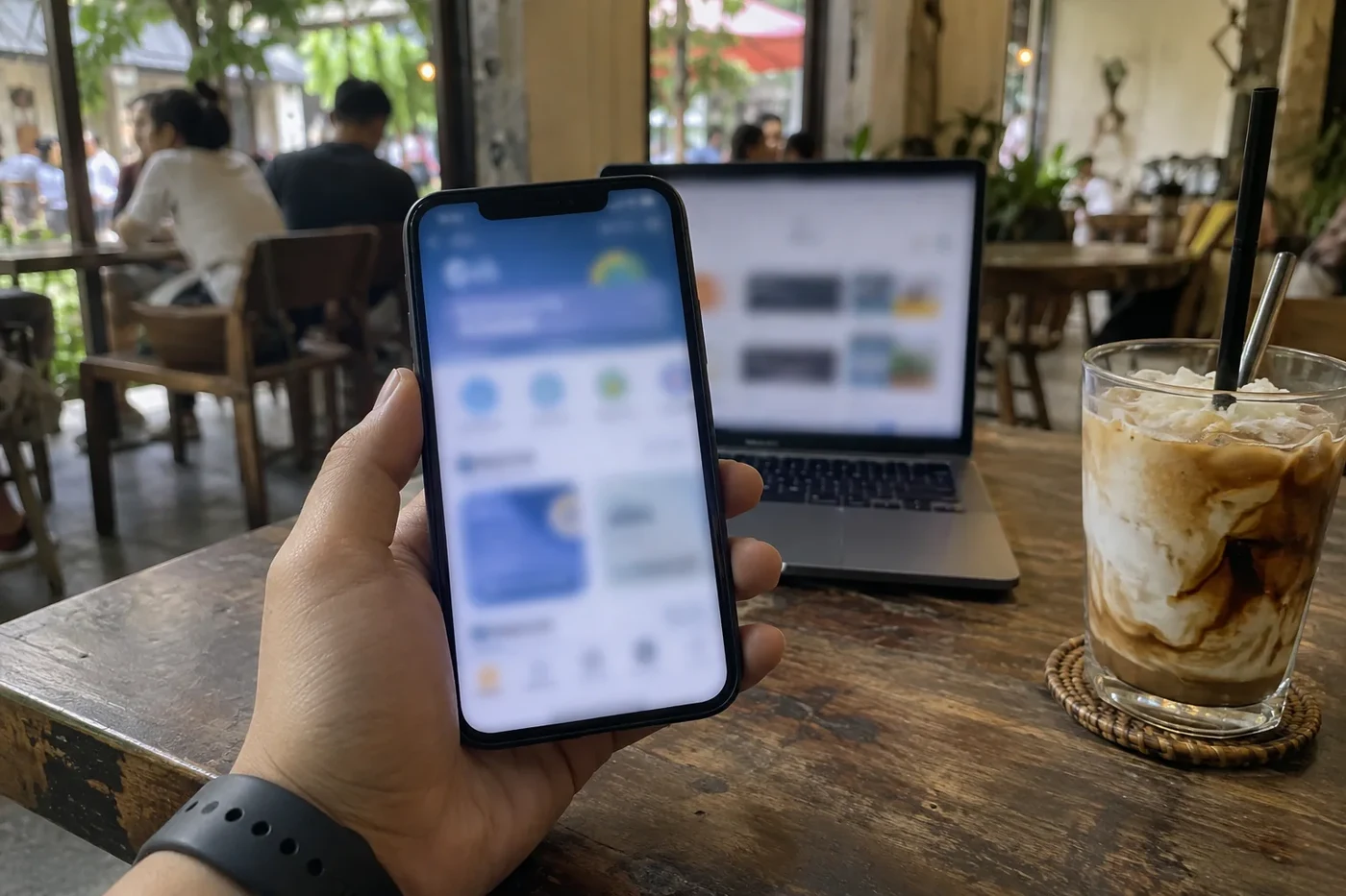 Hand holding a smartphone with an app open next to a laptop and an iced coffee in a cafe