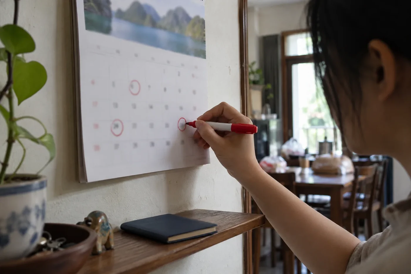 A woman circles dates on a wall calendar with a red marker, a passport lies on the shelf below