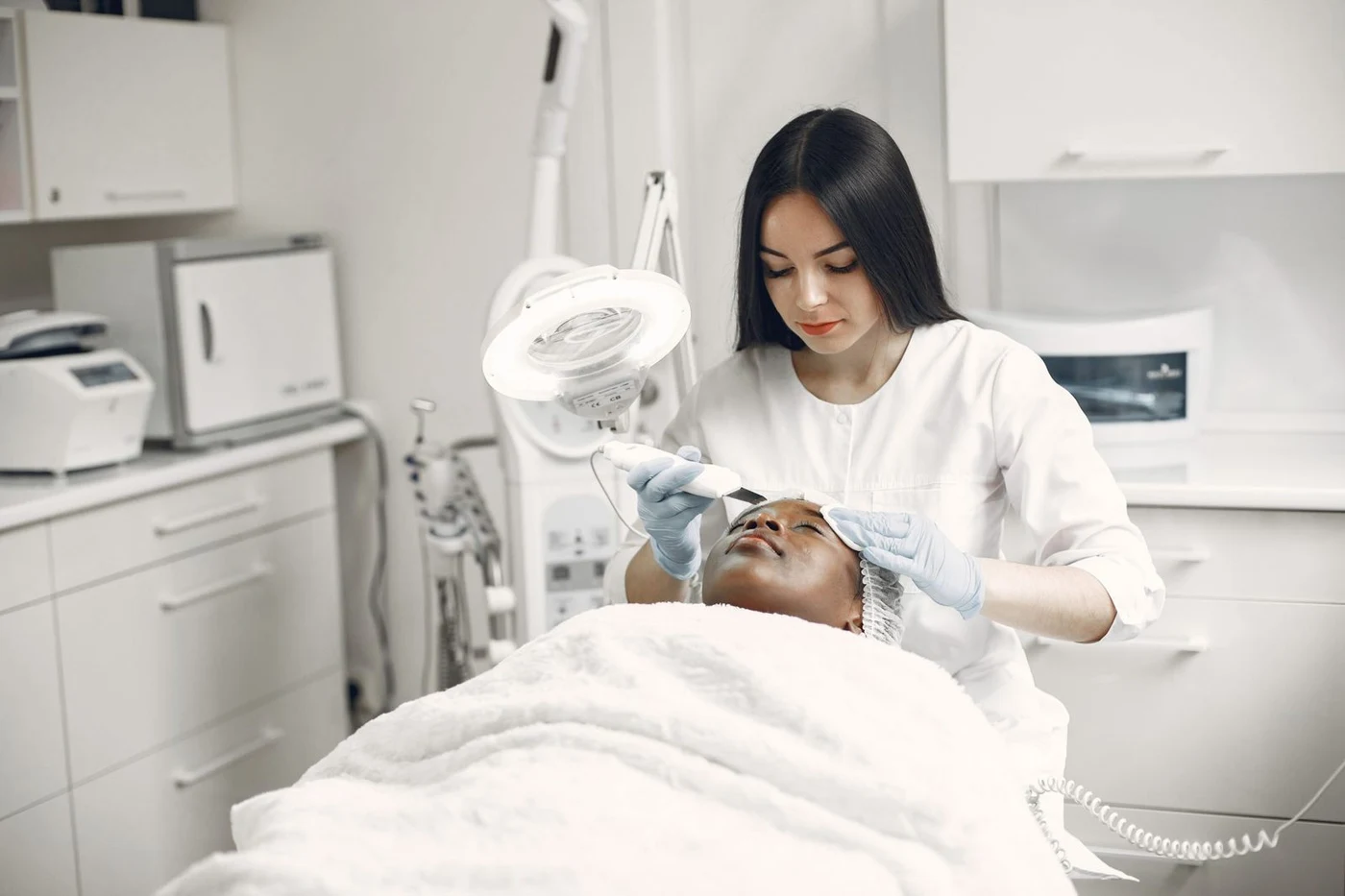 Cosmetologist in white wearing gloves treating a client’s face in a treatment room